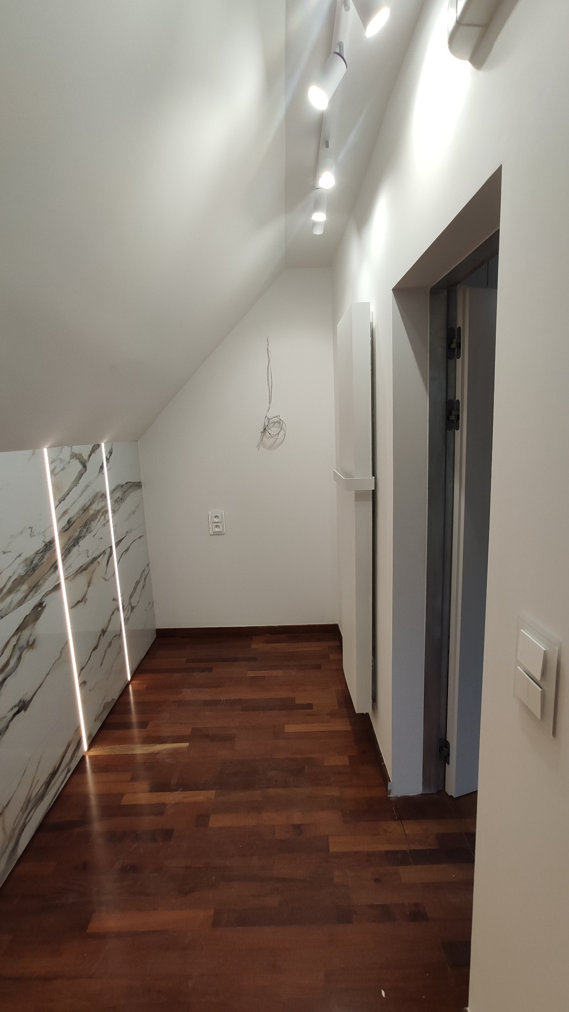 Wykończone poddasze ze ścianą z marmurową okładziną z podświetleniem LED, ciemną drewnianą podłogą, białymi ścianami i sufitem ze spotami oraz otwartymi szarymi drzwiami.