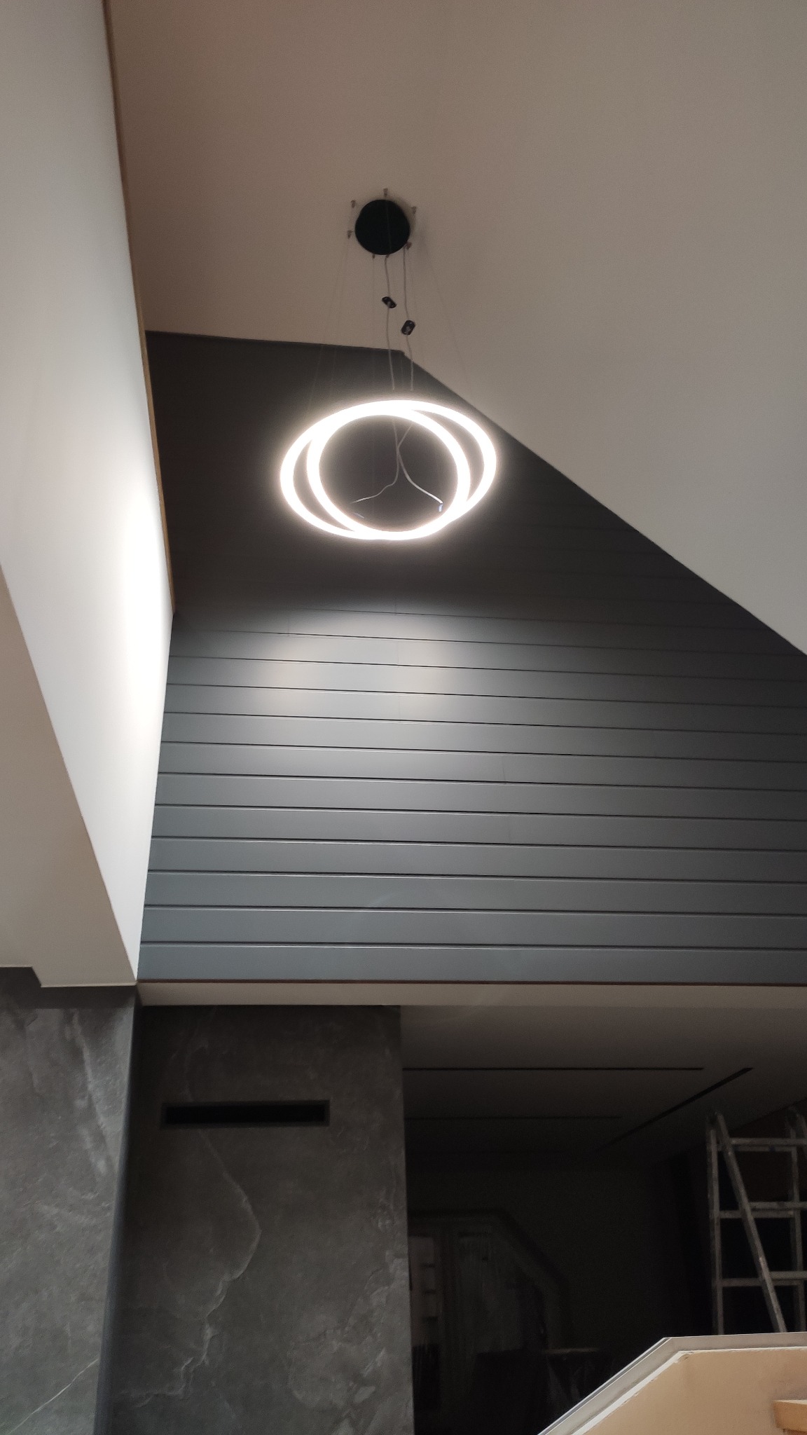 Nowoczesne wnętrze w Katowicach z podświetlanym, okrągłym żyrandolem LED na tle szarej, panelowej ściany i fragmentu marmurowej kolumny. Widoczny fragment drabiny w głębi pomieszczenia.