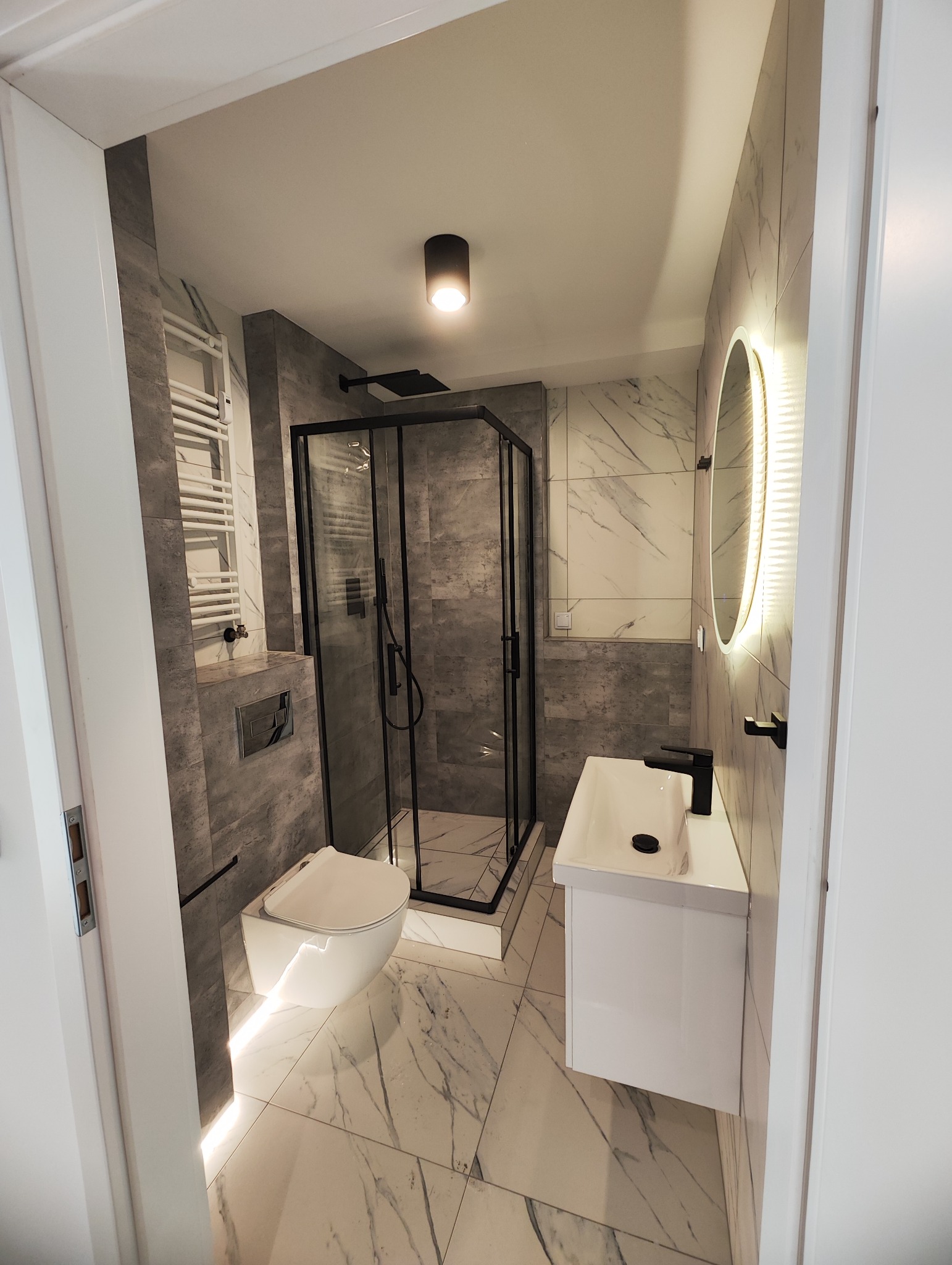 Nowoczesna łazienka z kabiną prysznicową z czarnymi ramami, podświetlanym lustrem, wiszącą toaletą i marmurowymi płytkami na podłodze i ścianach.