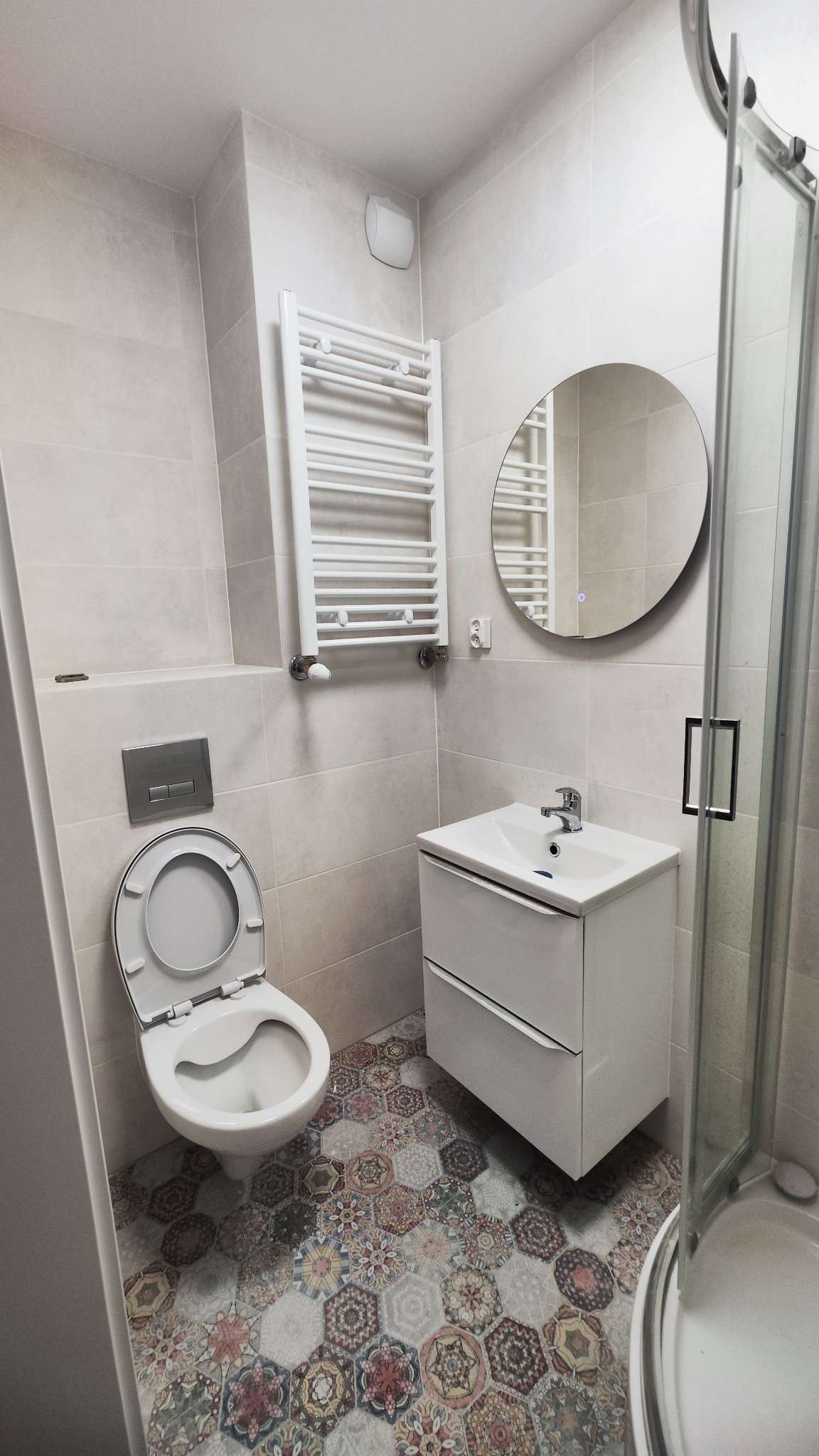 Wykończona łazienka z płytkami heksagonalnymi w stylu vintage na podłodze, białą ceramiką i grzejnikiem drabinkowym, okrągłe lustro z podświetleniem LED, kabina prysznicowa z transparentnego szkła.