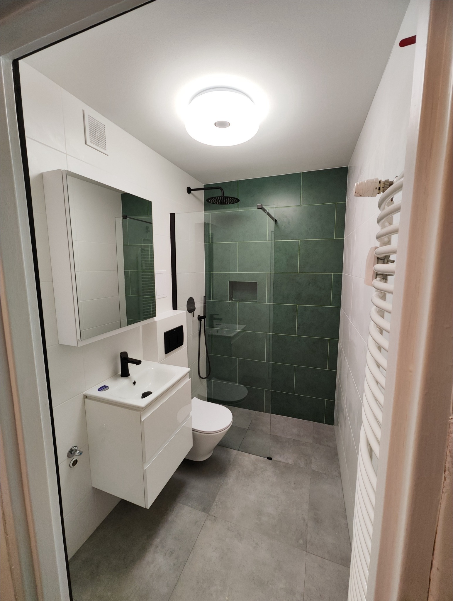 Nowoczesna łazienka po renowacji z kabiną prysznicową typu walk-in, zielonymi płytkami i białą armaturą z czarnymi akcentami, widoczny grzejnik drabinkowy.