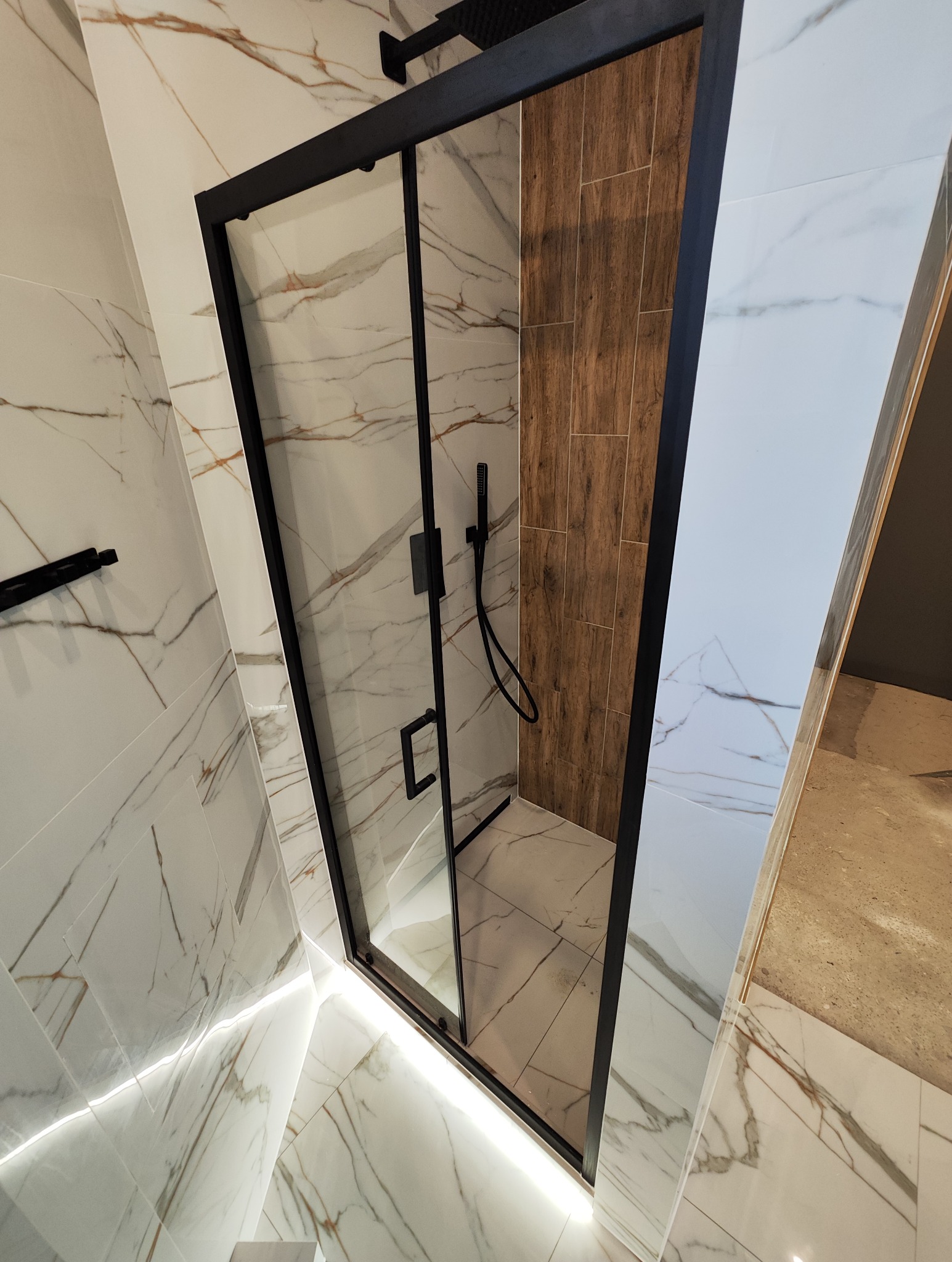Nowoczesna kabina prysznicowa z czarną ramą i szklanymi drzwiami, otoczona białymi płytkami imitującymi marmur z delikatnym podświetleniem LED u dołu, oraz ścianą wykończoną płytkami drewnopodobnymi.