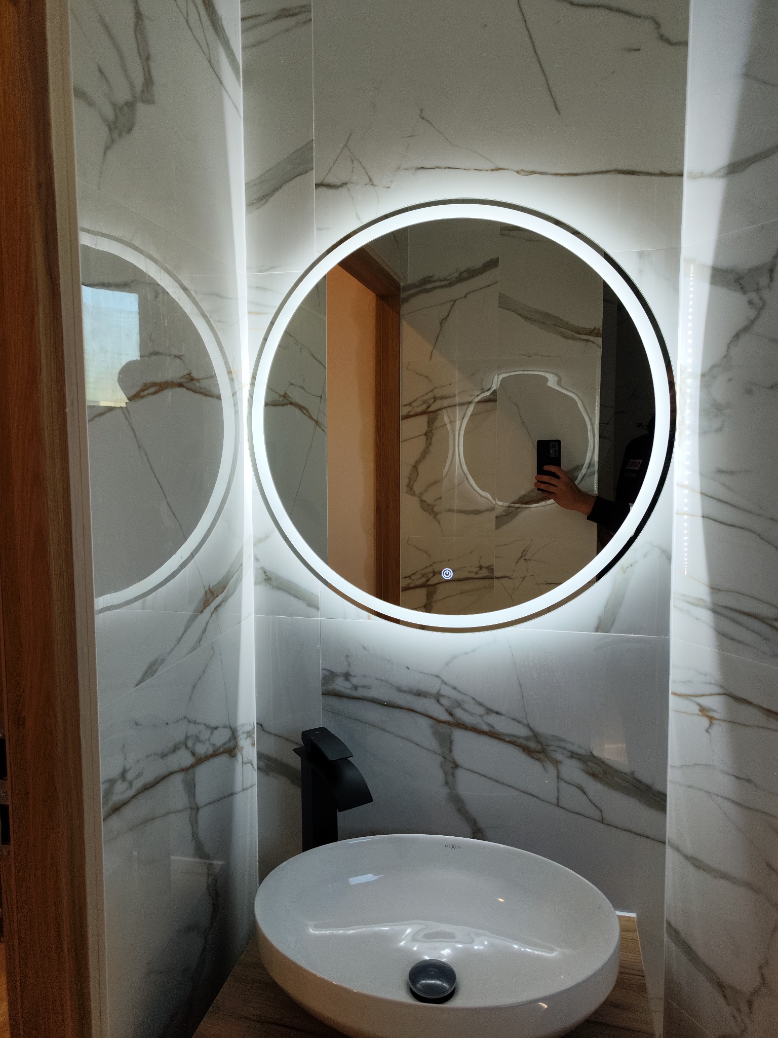 Nowoczesna łazienka z okrągłym lustrem podświetlanym LED, marmurowe płytki na ścianach, biała umywalka nablatowa i czarna bateria.