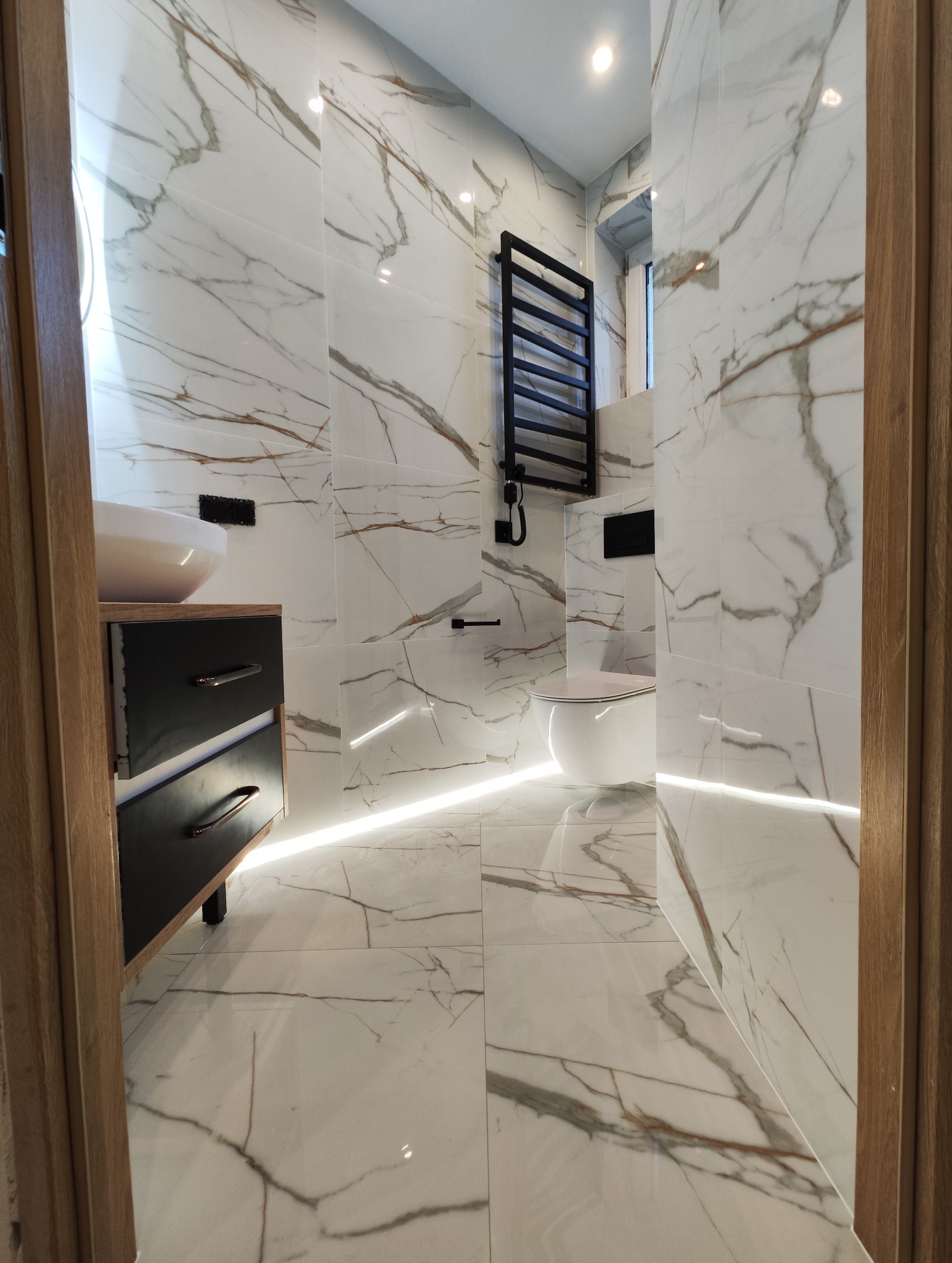 Nowoczesna łazienka z białymi płytkami imitującymi marmur, podświetleniem LED przy podłodze, czarnym grzejnikiem drabinkowym i drewnianą ramą drzwi w tle.