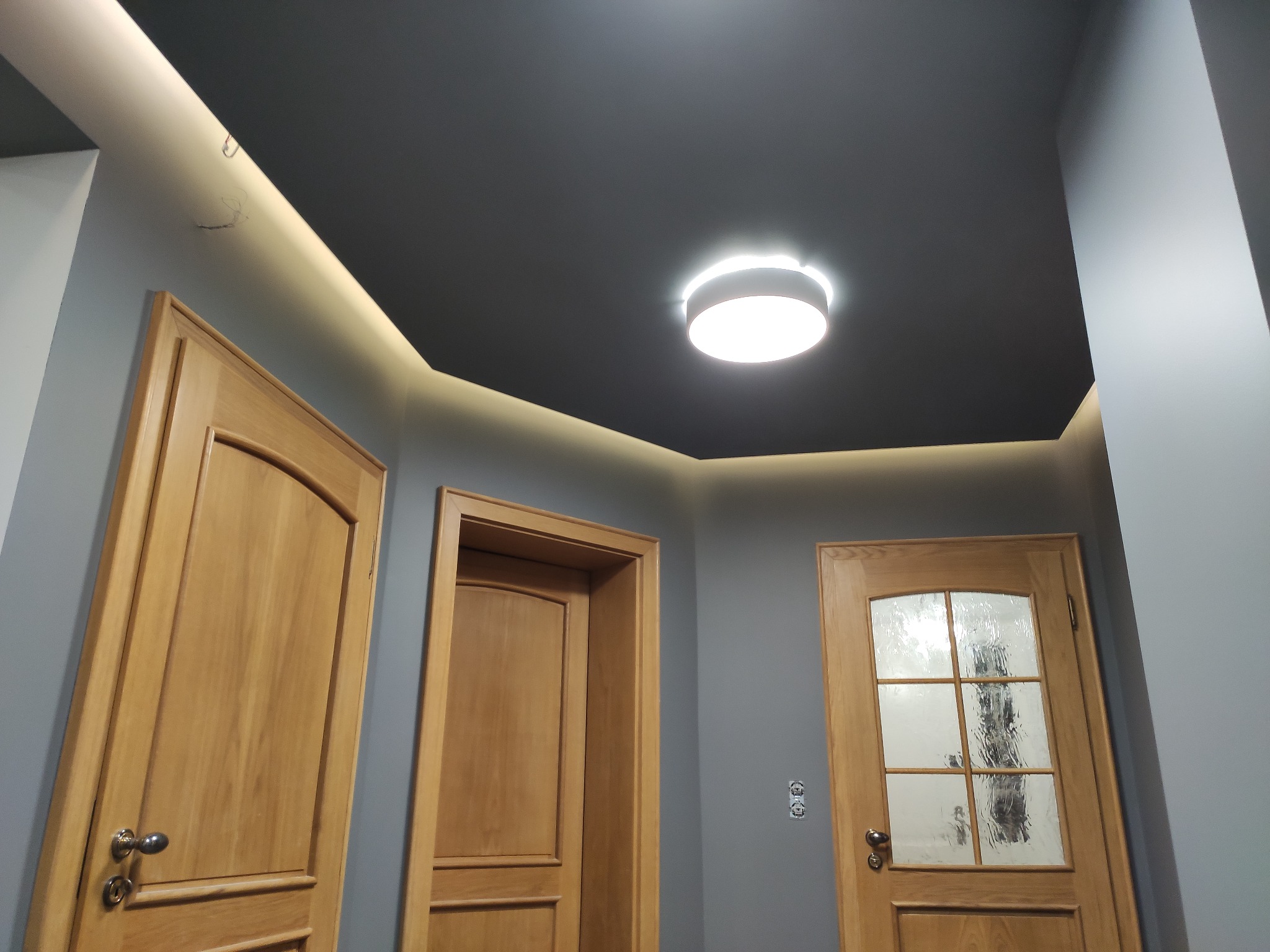 Wnętrze korytarza z trzema drewnianymi drzwiami, szarą ścianą i ciemnym sufitem z okrągłą lampą oraz oświetleniem LED wzdłuż krawędzi.