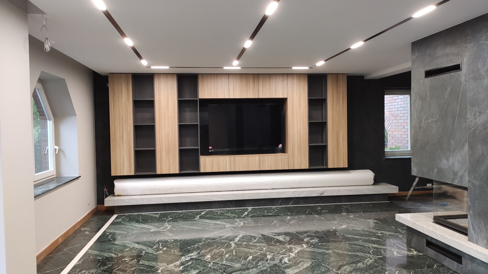 Nowoczesny salon z marmurową podłogą i zabudową telewizyjną z półkami, oświetlenie szynowe na suficie.