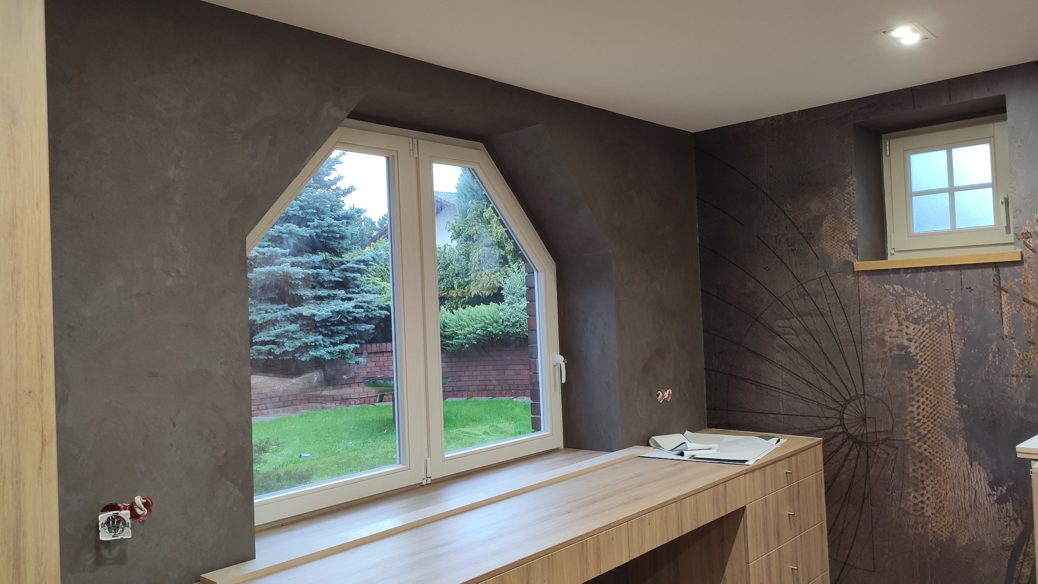 Pokój na poddaszu z trapezoidalnym oknem, szarą ścianą o fakturze betonu, drewnianym blatem pod oknem i tapetą w stylu vintage z motywem koła na przeciwległej ścianie.