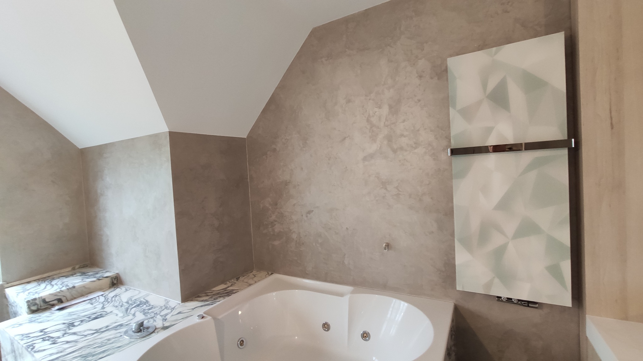 Elegancka łazienka na poddaszu z wanną jacuzzi, ściany wykończone dekoracyjnym tynkiem w odcieniach szarości, biała armatura i grzejnik panelowy z geometrycznym wzorem.
