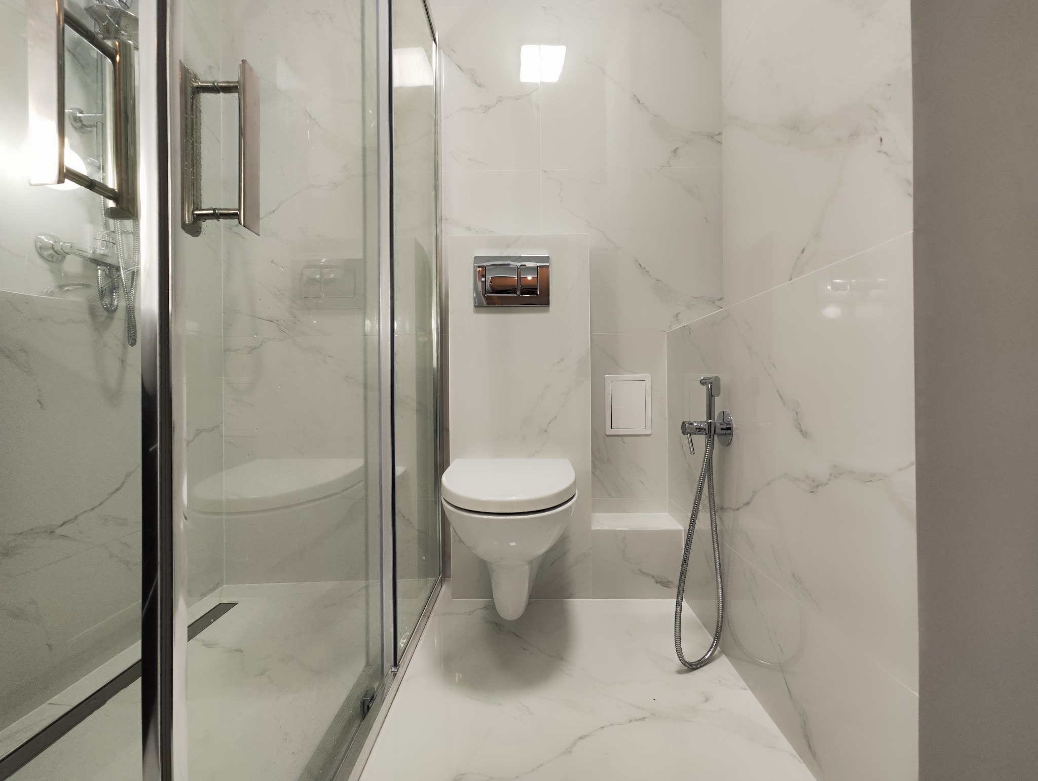 Nowoczesna łazienka z białymi płytkami imitującymi marmur, wisząca toaleta z chromowanym przyciskiem spłukiwania, szklana kabina prysznicowa z chromowanymi okuciami i bidetka.