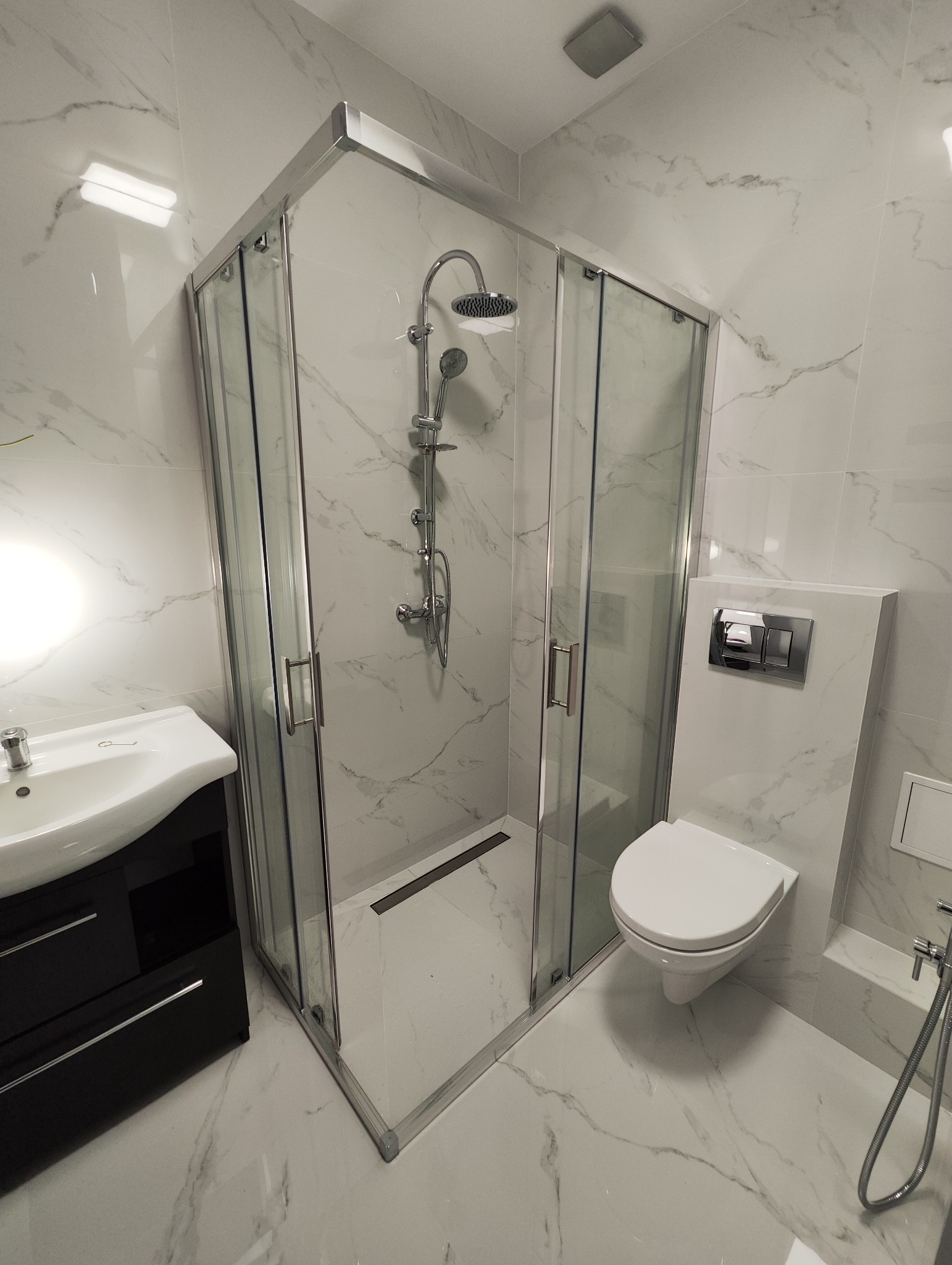 Nowoczesna łazienka z kabiną prysznicową z chromowanymi profilami, umywalką z szafką w kolorze czarnym i podwieszaną toaletą, wykończona białymi płytkami imitującymi marmur.