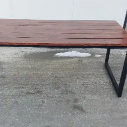 Stół z ciemnobrązowym blatem z desek i czarnymi, metalowymi nogami o trapezowym kształcie, ustawiony na betonowej powierzchni z resztkami śniegu.