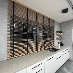 Jasne drewniane żaluzje poziome z czarnymi sznurkami w nowoczesnej kuchni z szarą ścianą, białymi szafkami i szarym blatem