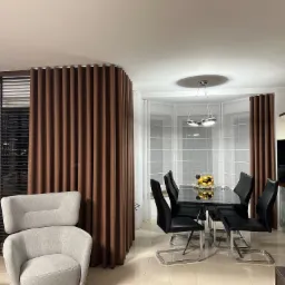 Nowoczesne wnętrze z brązowymi, falowanymi zasłonami oddzielającymi strefę wypoczynkową z szarym fotelem od jadalni z czarnym stołem i krzesłami.