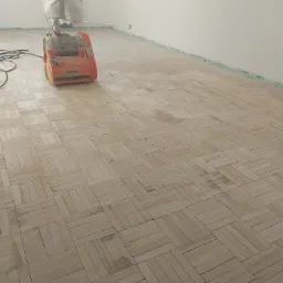 Parkiet w jodełkę podczas cyklinowania maszyną Scorpion, widoczne ślady po szlifowaniu i taśma malarska zabezpieczająca ściany.