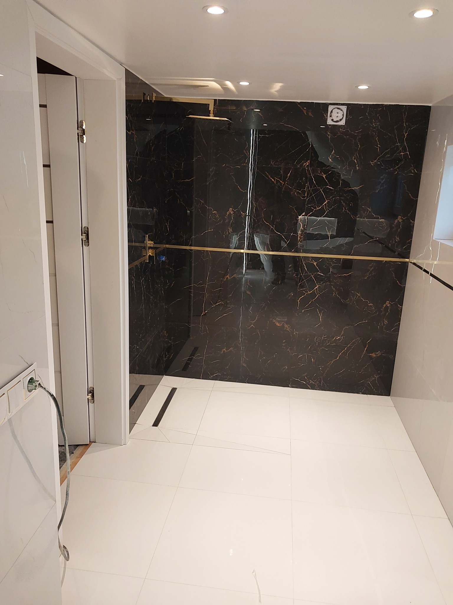 Nowoczesna łazienka z czarnymi płytkami imitującymi marmur, złotymi akcentami w kabinie prysznicowej i białymi płytkami podłogowymi, widok na otwartą kabinę prysznicową.