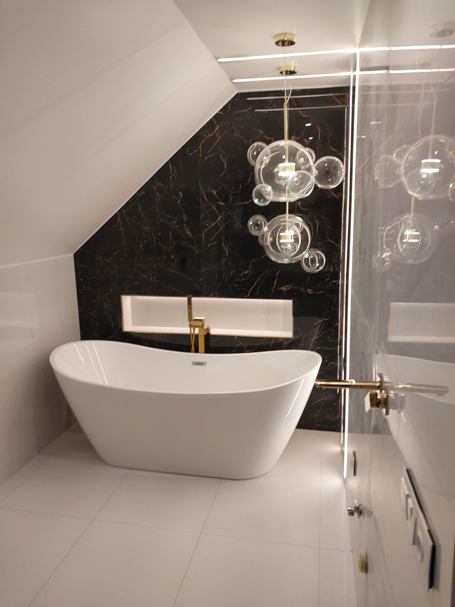 Nowoczesna łazienka na poddaszu z wolnostojącą białą wanną, armaturą w kolorze złotym i dekoracyjną lampą z szklanych kul.