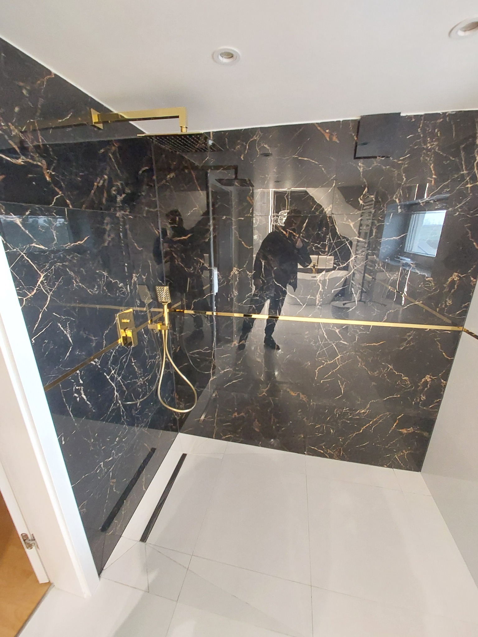 Nowoczesny prysznic walk-in z czarnymi płytkami imitującymi marmur z złotymi żyłami, biała podłoga z płytek, złota armatura i szklana ścianka. Widoczne odbicie osoby robiącej zdjęcie.