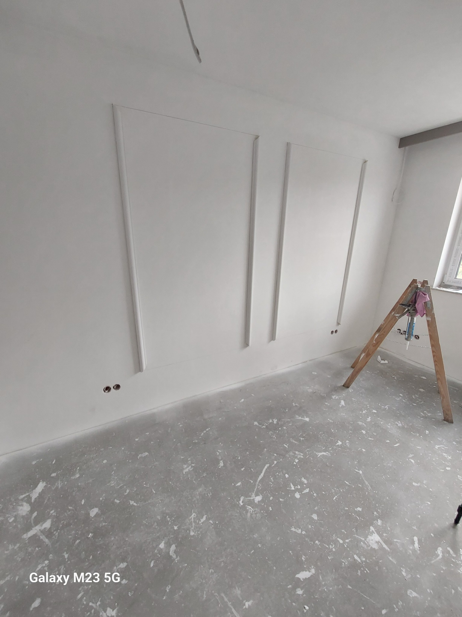 Świeżo pomalowane, białe ściany z dekoracyjnymi listwami w Tarnobrzegu, widoczna drabina malarska i ślady farby na podłodze.