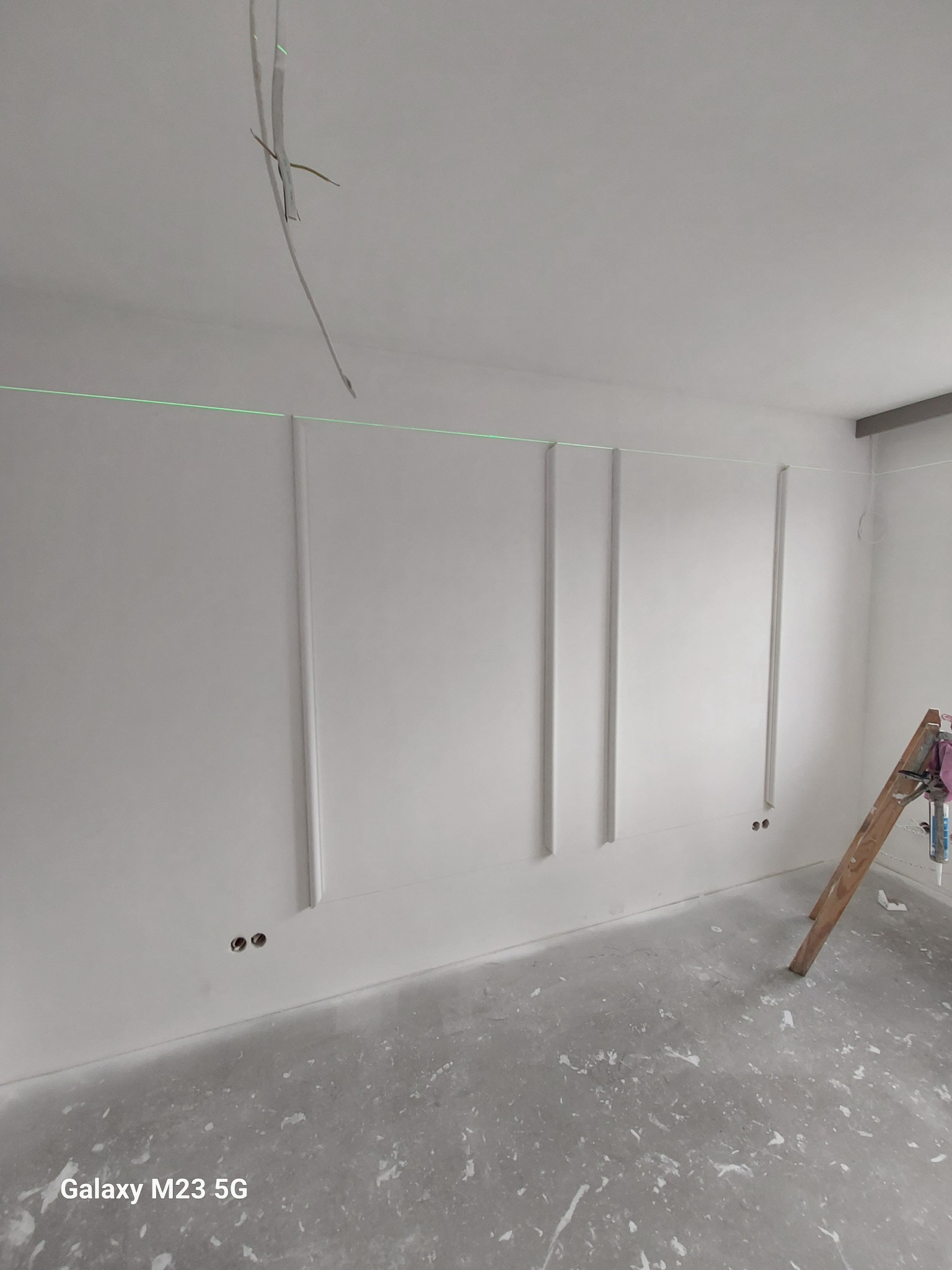 Białe ściany z dekoracyjnymi listwami w trakcie montażu, widoczna linia lasera do poziomowania, drabina oparta o ścianę, surowa podłoga z resztkami zaprawy.