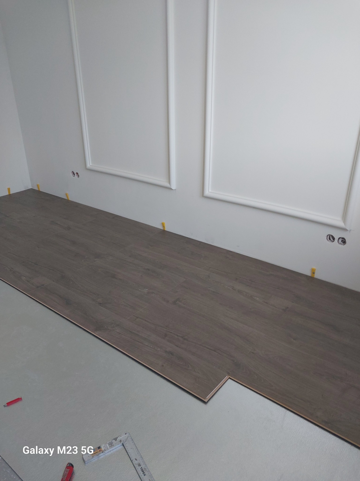 Układanie paneli podłogowych w pomieszczeniu z białymi ścianami i dekoracyjnymi listwami; widoczne narzędzia i żółte kliny dystansowe.