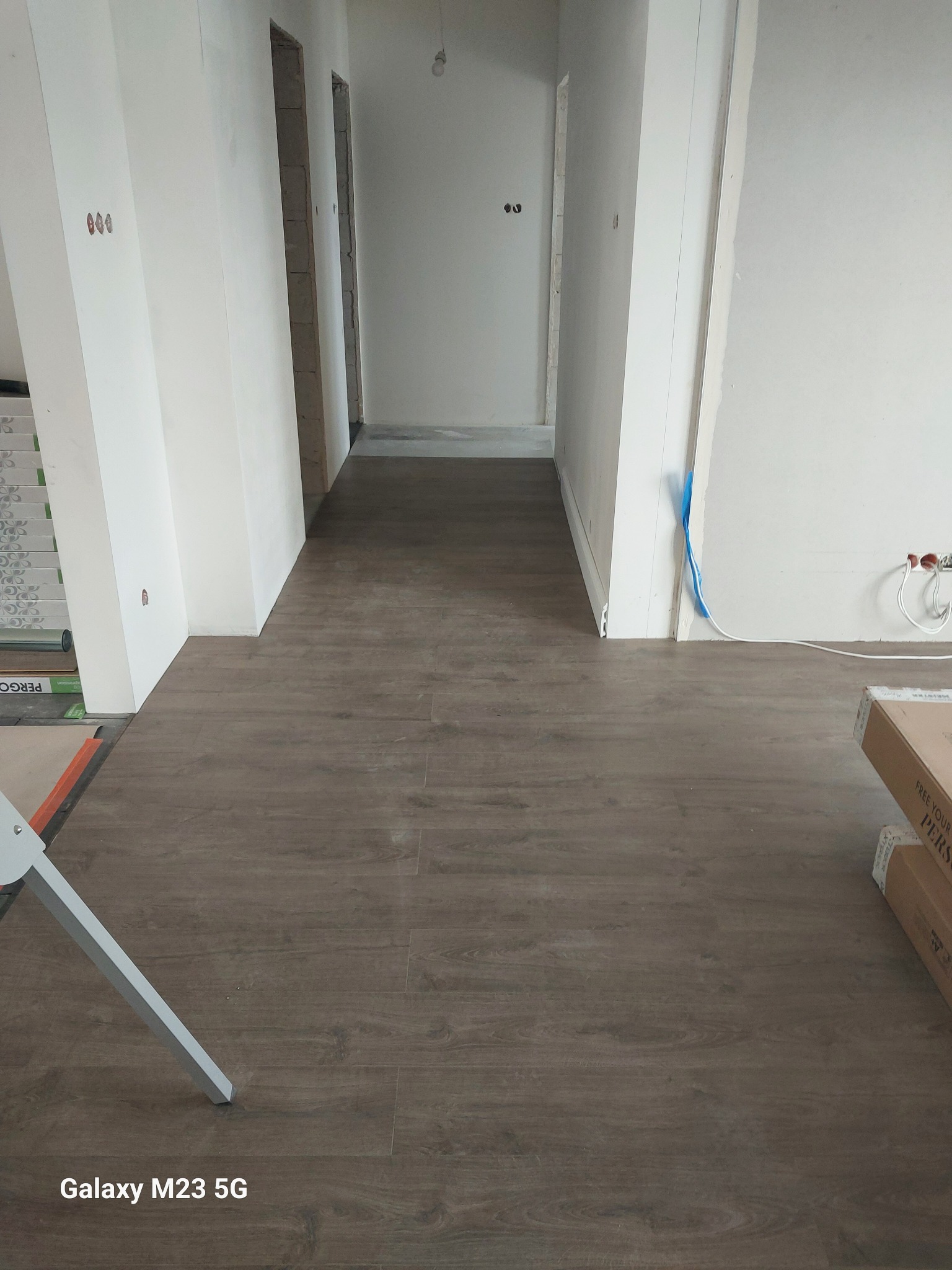 Nowo położone panele podłogowe w korytarzu, widoczne puste otwory drzwiowe i puszki elektryczne, częściowo zakryte opakowania paneli i noga drabiny malarskiej.