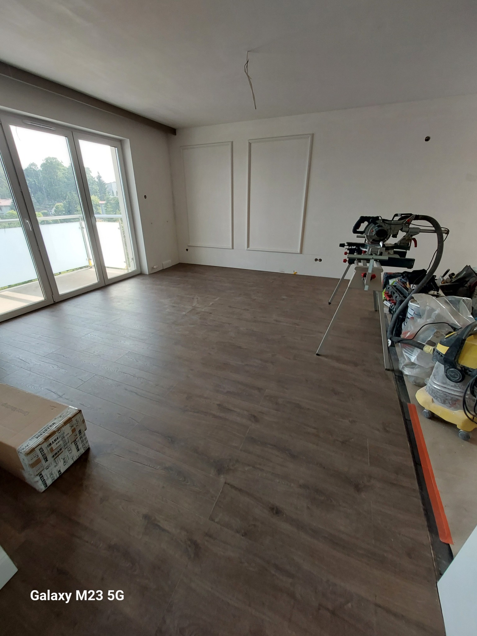 Wnętrze pokoju z nowo ułożonymi panelami podłogowymi w odcieniach brązu, widoczne narzędzia pracy, w tym piła ukośnica na statywie i odkurzacz przemysłowy, karton z panelami oraz duże okno...