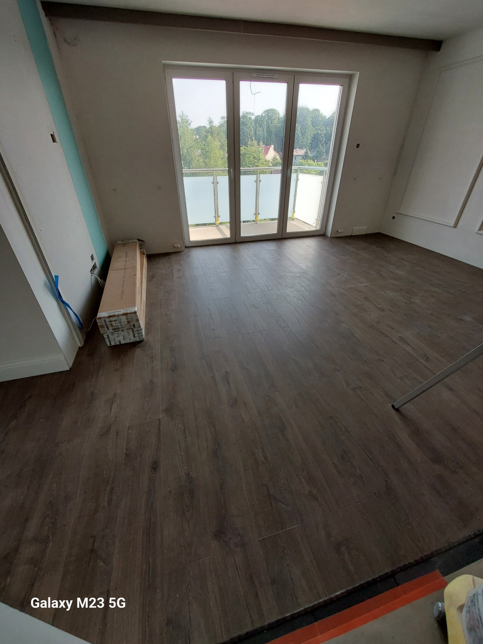 Ułożone panele podłogowe w pokoju z wyjściem na balkon, widoczne kartony paneli przy ścianie, częściowo widoczne narzędzia w rogu.