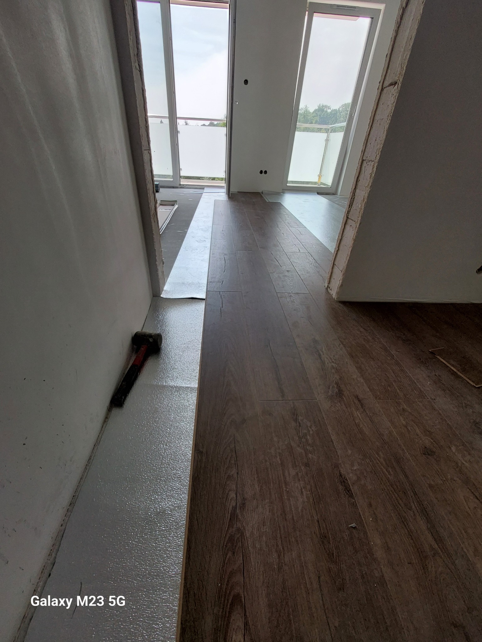 Montaż paneli podłogowych w jasnym pomieszczeniu, widoczny młotek przy ścianie, część podłogi wyłożona folią izolacyjną.