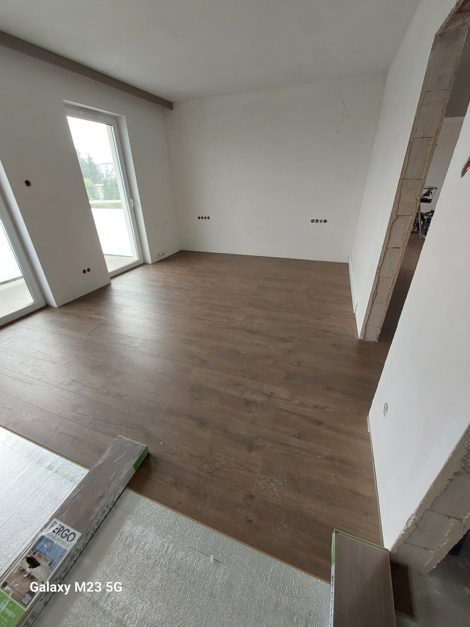 Wnętrze pokoju z częściowo ułożoną podłogą z paneli laminowanych w odcieniach brązu, widoczne opakowanie paneli marki Fergo oraz podkład pod panele, ściany pomalowane na biało, surowy otwór drzwiowy.