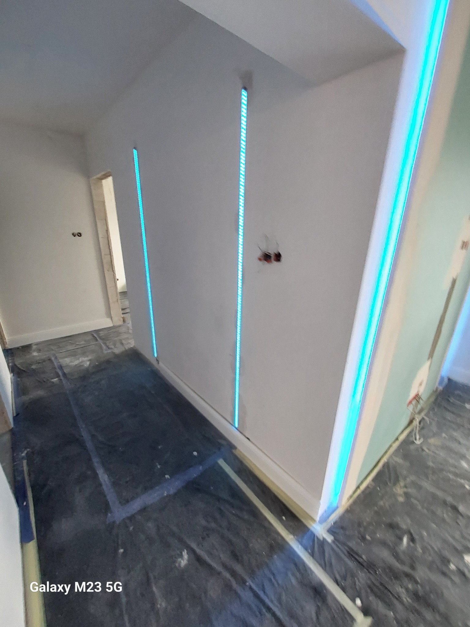 Korytarz w trakcie remontu z zamontowanymi pionowymi listwami LED w ścianach, zabezpieczona folią podłoga i widoczne kable elektryczne.