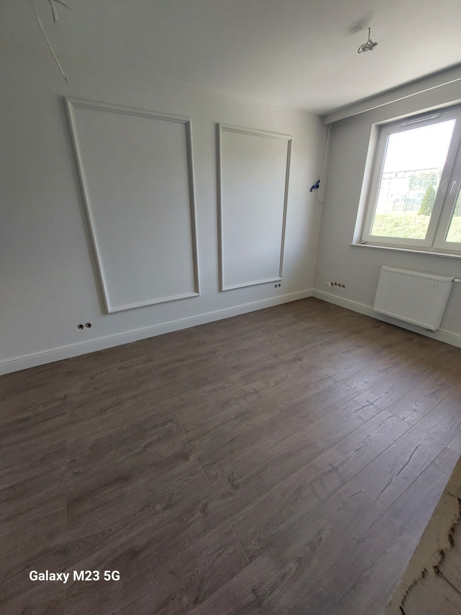 Jasne wnętrze po remoncie z panelami podłogowymi w odcieniach brązu, białymi ścianami z dekoracyjnymi listwami, grzejnikiem pod oknem i wystającymi kablami ze ściany.