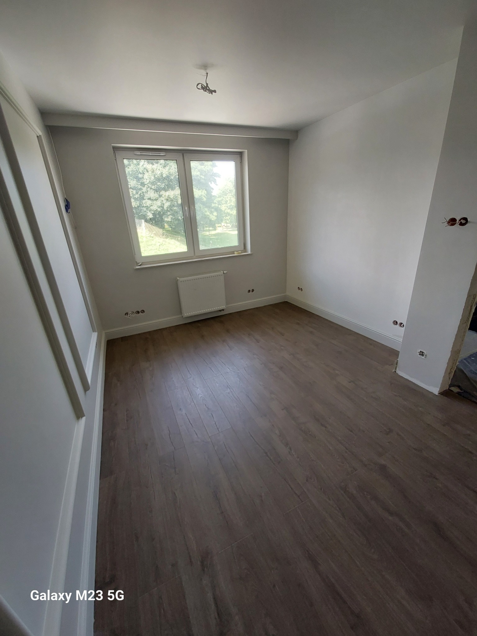 Pusty pokój po remoncie z brązowymi panelami podłogowymi, białymi ścianami z widocznymi gniazdkami elektrycznymi i grzejnikiem pod oknem.