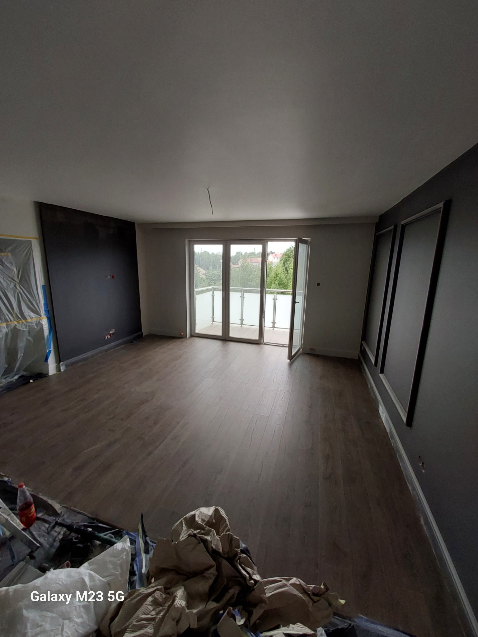 Pokój po remoncie z panelami podłogowymi, czarną ścianą i dekoracyjnymi listwami, widok na balkon z otwartymi drzwiami, ślady prac wykończeniowych widoczne w dolnym rogu.