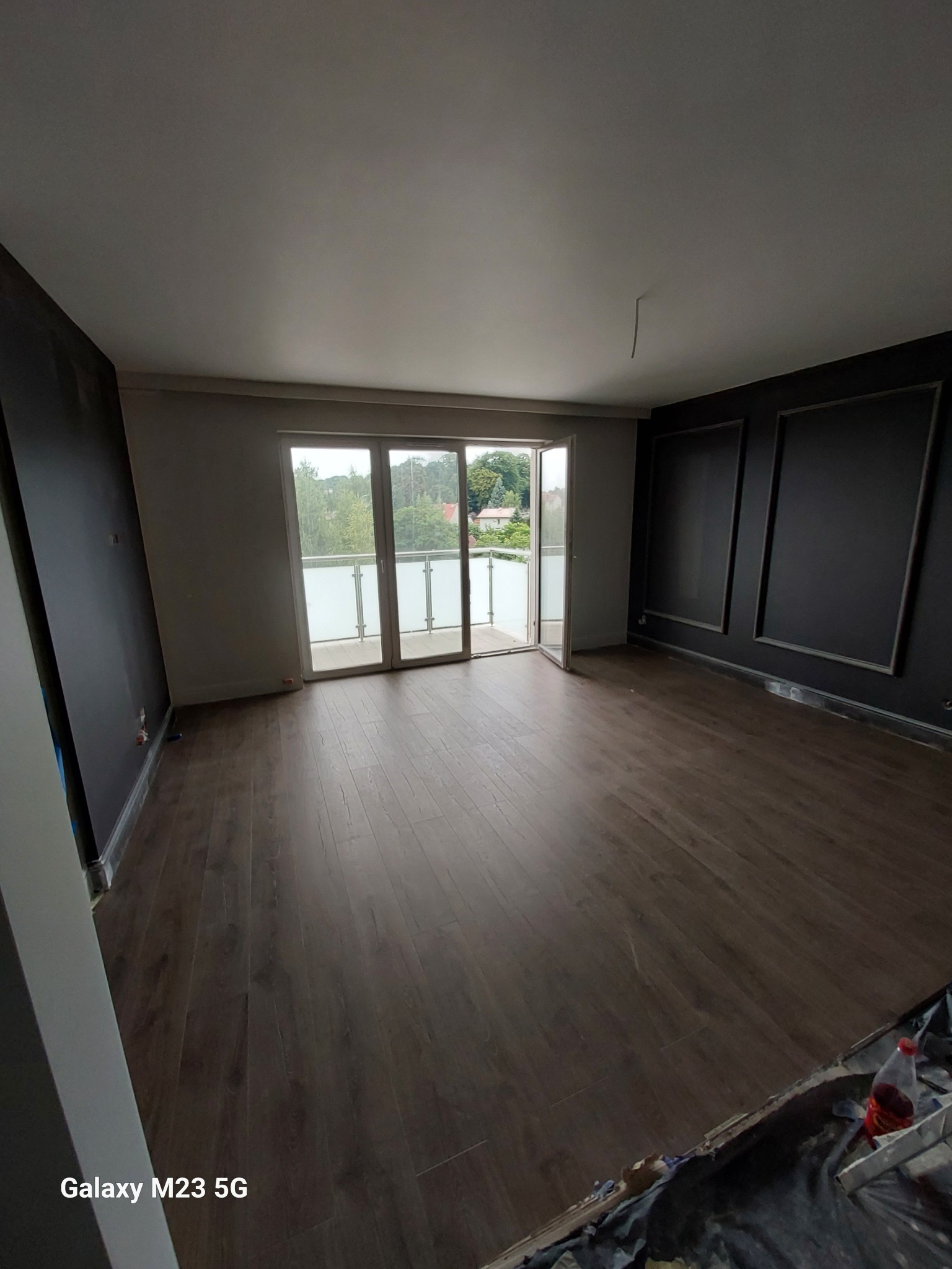 Pokój po remoncie z ciemnymi panelami podłogowymi i czarnymi ścianami z dekoracyjnymi ramkami, widok na balkon i zieleń za oknem, ślady prac wykończeniowych widoczne przy podłodze.