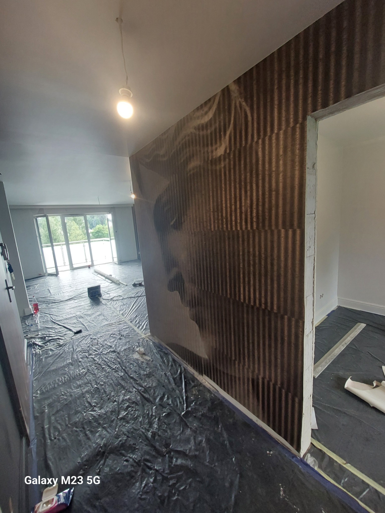 Wnętrze w trakcie prac wykończeniowych z widoczną ścianą z fototapetą z motywem twarzy, podłoga zabezpieczona folią malarską, widoczne narzędzia i materiały budowlane.