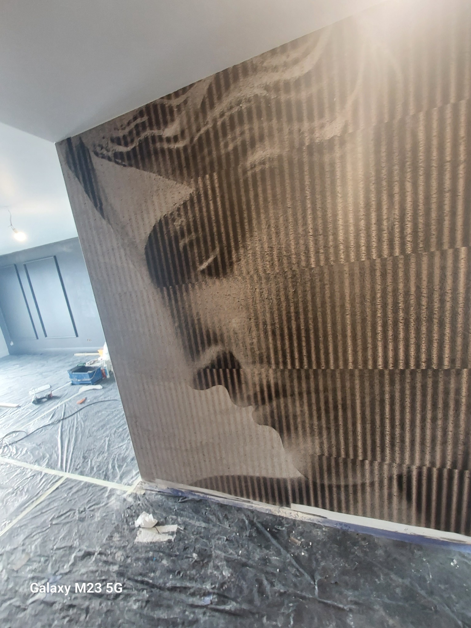 Ściana z abstrakcyjnym wzorem przypominającym twarz, zabezpieczona folią podłoga, widoczne narzędzia malarskie w tle.