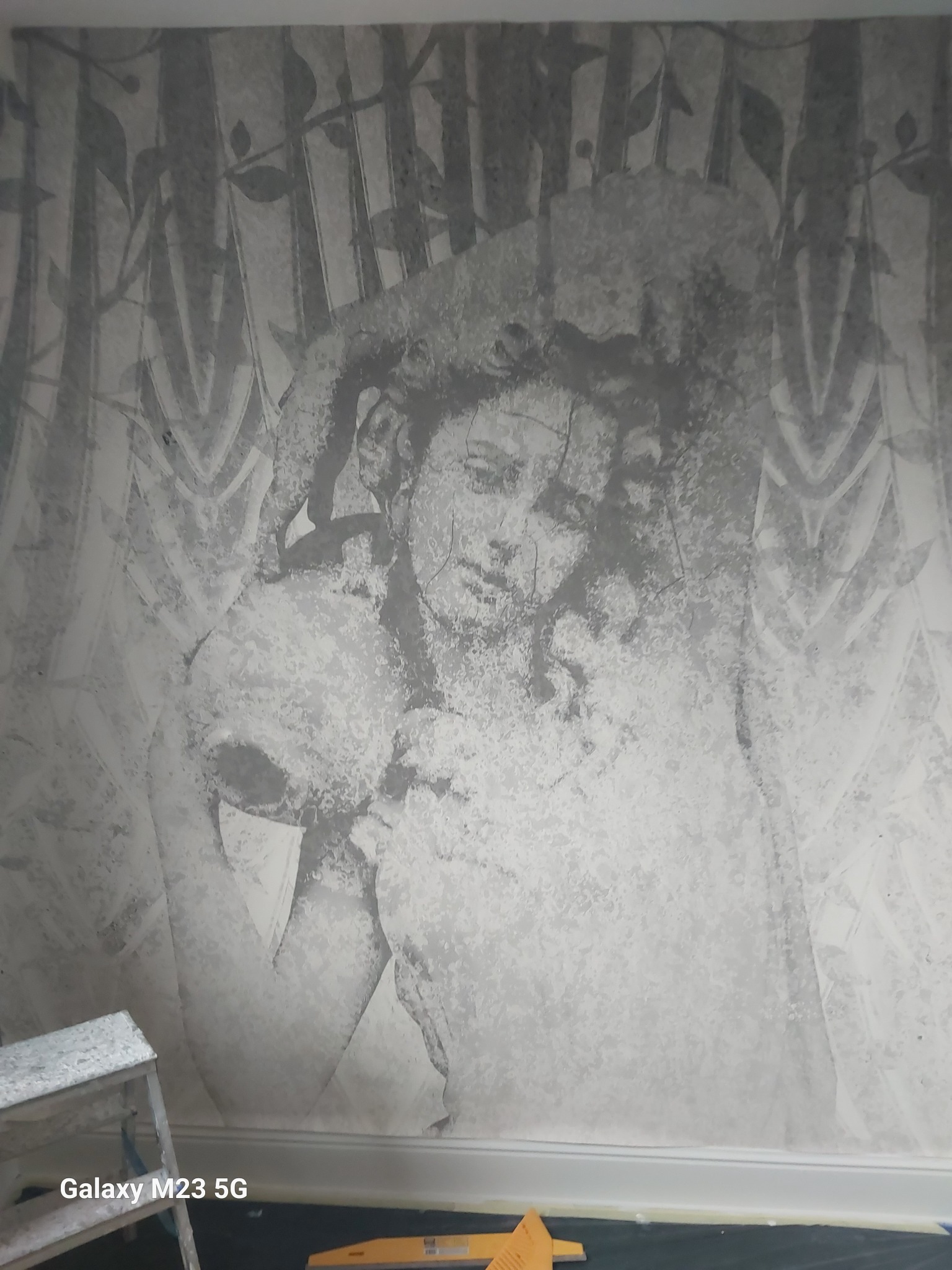 Ściana z szarym, artystycznym malunkiem przedstawiającym postać kobiety w kapeluszu na tle stylizowanych drzew, widoczny fragment drabiny malarskiej i poziomicy na podłodze.