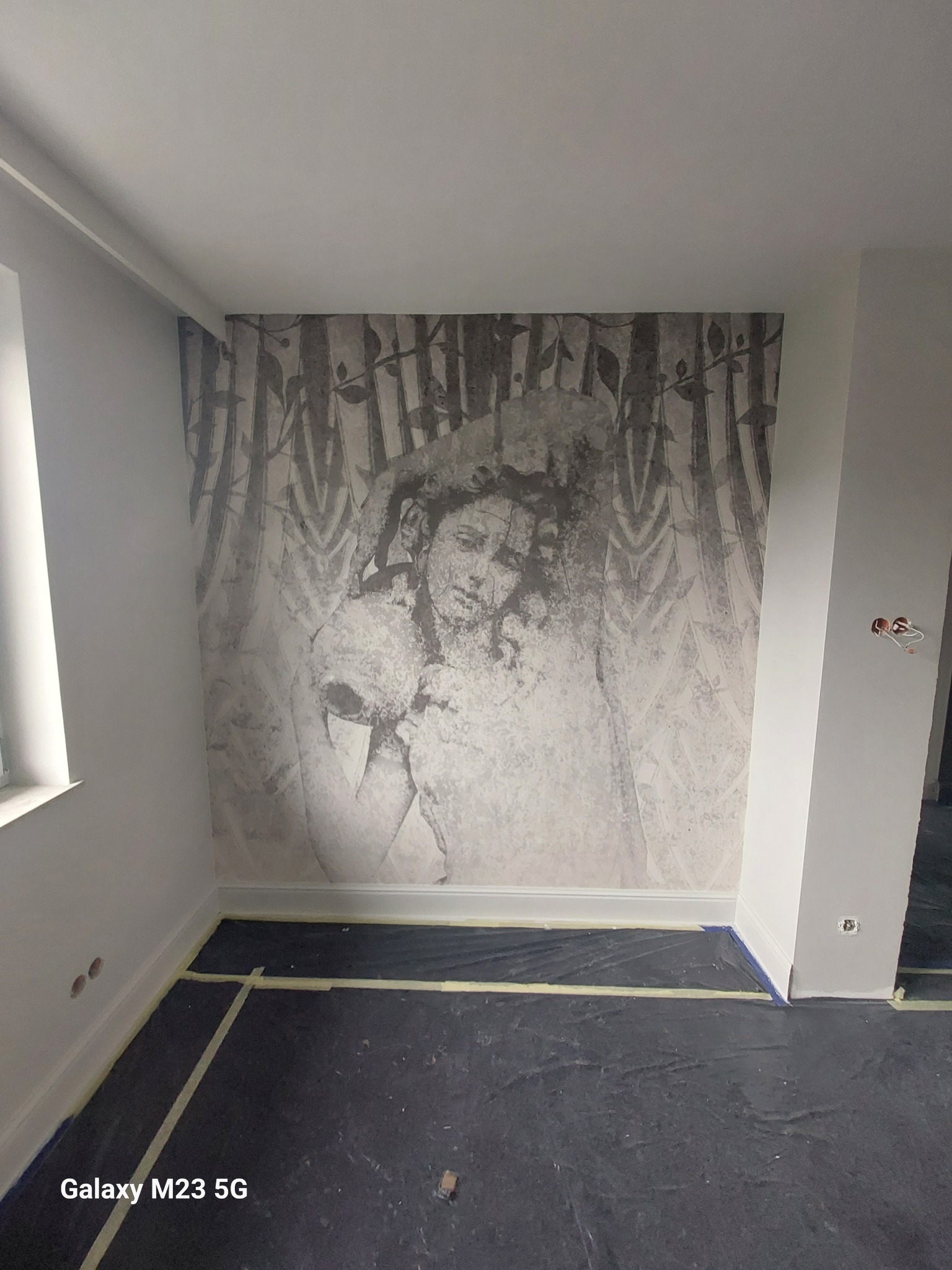 Pokój z szarą fototapetą przedstawiającą postać w stylu vintage, podłoga zabezpieczona folią malarską, widoczne puszki elektryczne w ścianie.