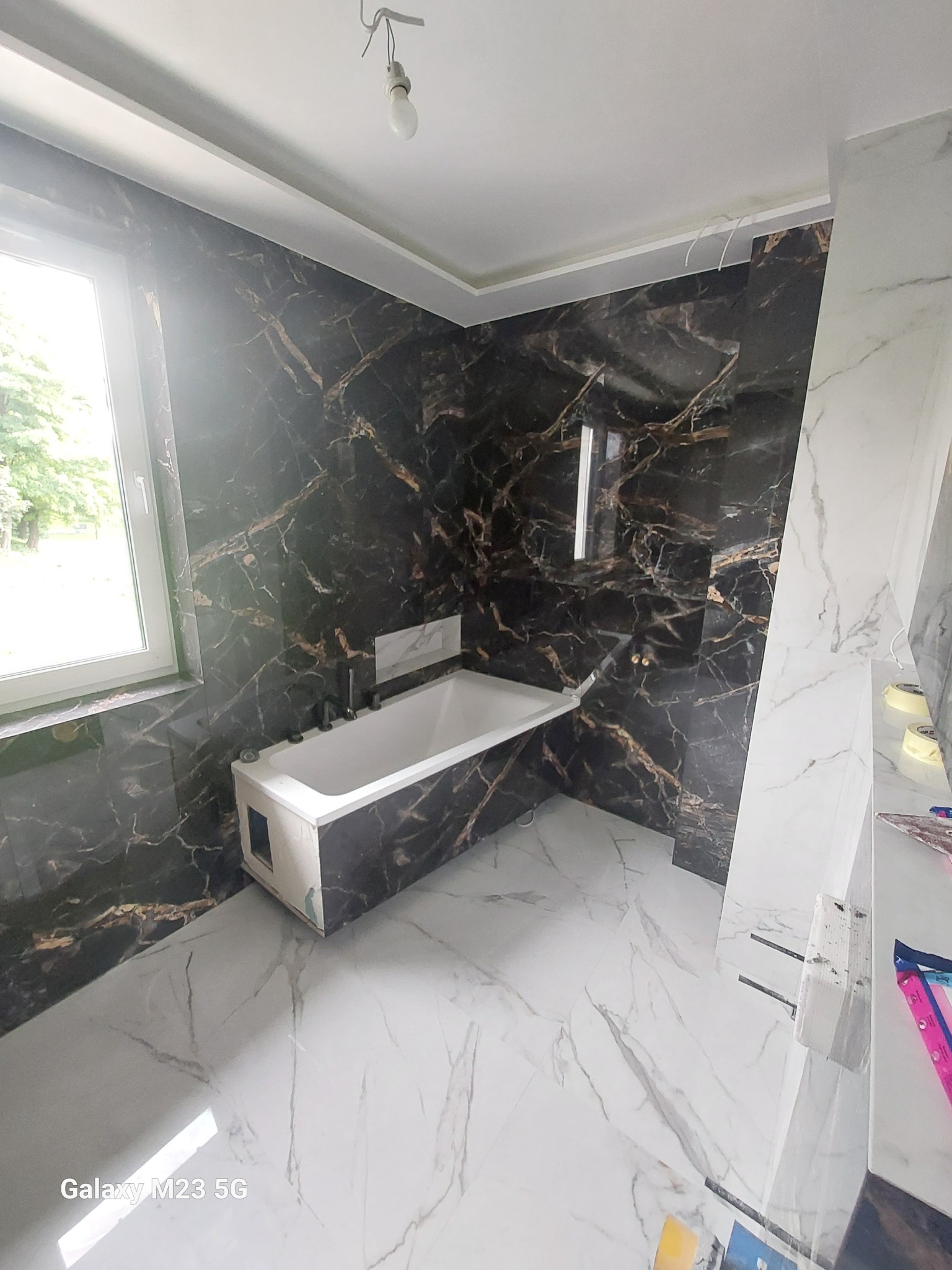 Wnętrze łazienki w trakcie remontu: biała wanna częściowo obudowana, czarne ściany imitujące marmur z złotymi żyłami, białe marmurowe płytki podłogowe, widoczne narzędzia i materiały budowlane.