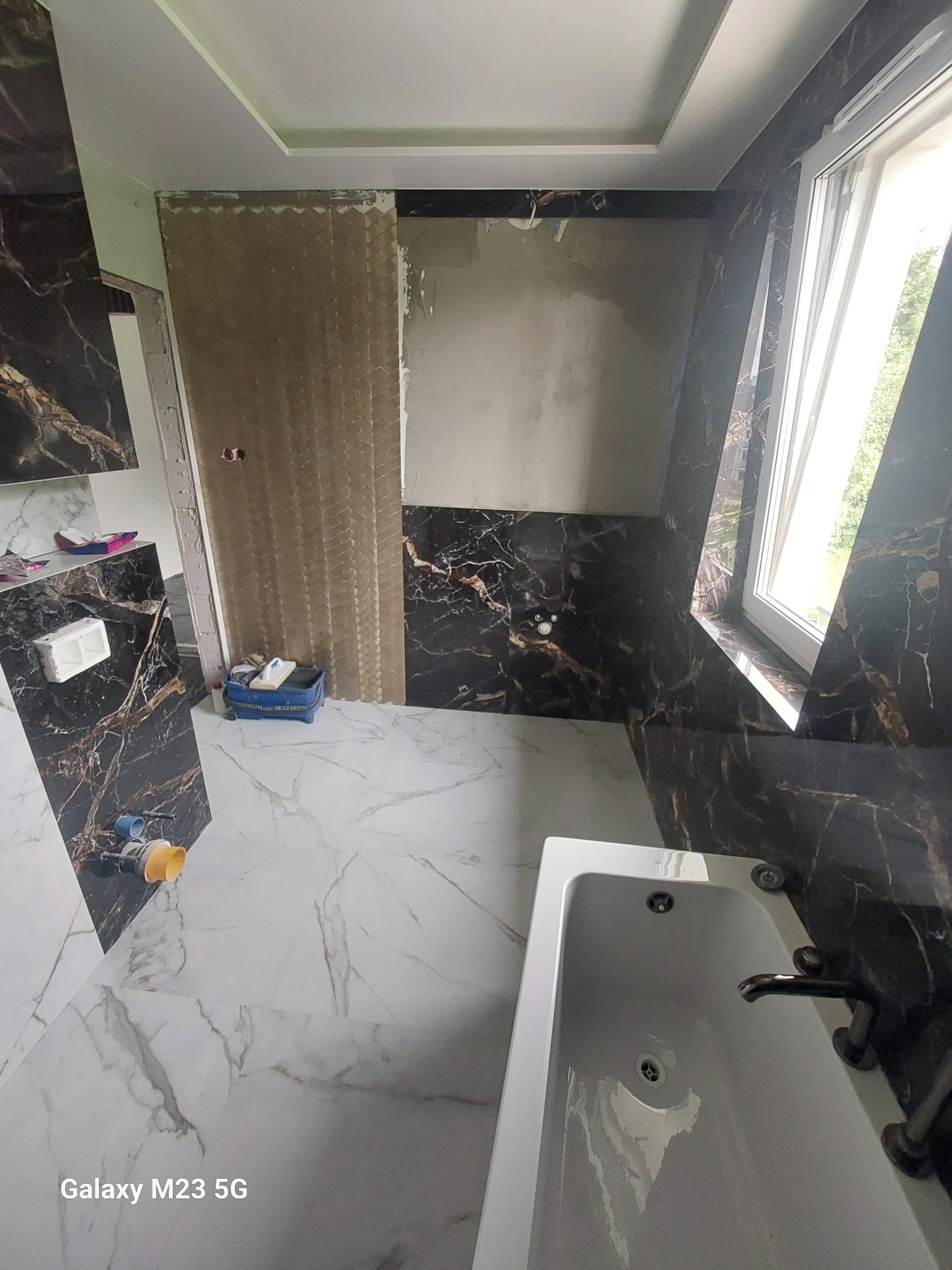 Wnętrze łazienki w trakcie remontu: częściowo wykończone ściany z czarnym marmurem, biała marmurowa podłoga, fragment wanny, fragment ściany przygotowany pod płytki z wzorem jodełki i narzędzia...