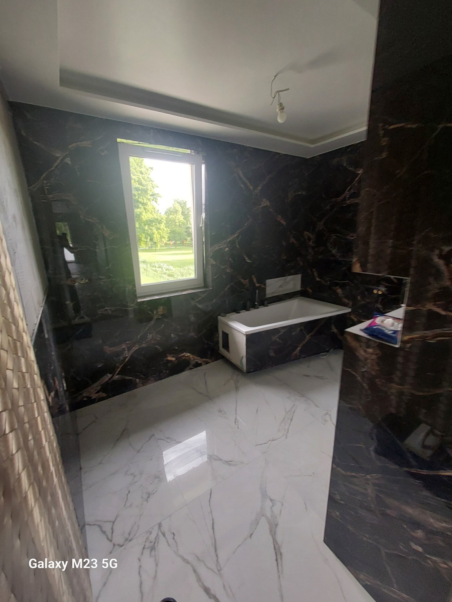 Wykończona łazienka z wanną, marmurowymi ścianami w odcieniach brązu i białą marmurową podłogą, widok na zieleń za oknem.