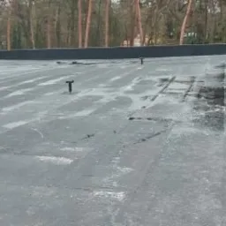 Płaski, pokryty bitumiczną papą dach po deszczu, z widocznymi elementami wentylacji i murkiem attykowym w tle.