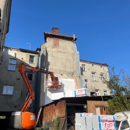 Rafał Rączy - Prace ociepleniowe na elewacji budynku wykonywane z podnośnika koszowego; widoczne płyty styropianowe z nadrukiem producenta, częściowo ocieplona ściana i pomarańczowy podnośnik.