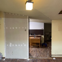 Rafał Rączy - Ściana z płyty gipsowo-kartonowej po szpachlowaniu, widoczne ślady po łatach, podłoga z parkietu pokryta pyłem, w tle widoczny fragment kuchni.