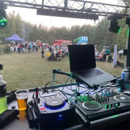 Rafał Rączy - Stanowisko DJ-a na imprezie plenerowej z widocznymi gramofonami, mikserem, laptopem, napojami i oświetleniem scenicznym, w tle bawiący się ludzie, wóz strażacki i dmuchana zjeżdżalnia.