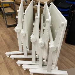 Składane, białe stoły konferencyjne na kółkach, ustawione jeden za drugim w magazynie, widoczne kartony i regał z narzędziami w tle.