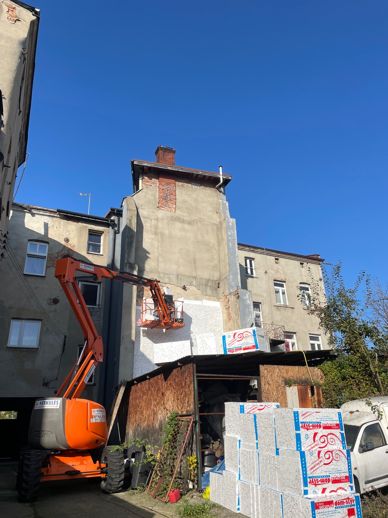 Prace ociepleniowe na elewacji budynku wykonywane z podnośnika koszowego; widoczne płyty styropianowe z nadrukiem producenta, częściowo ocieplona ściana i pomarańczowy podnośnik.