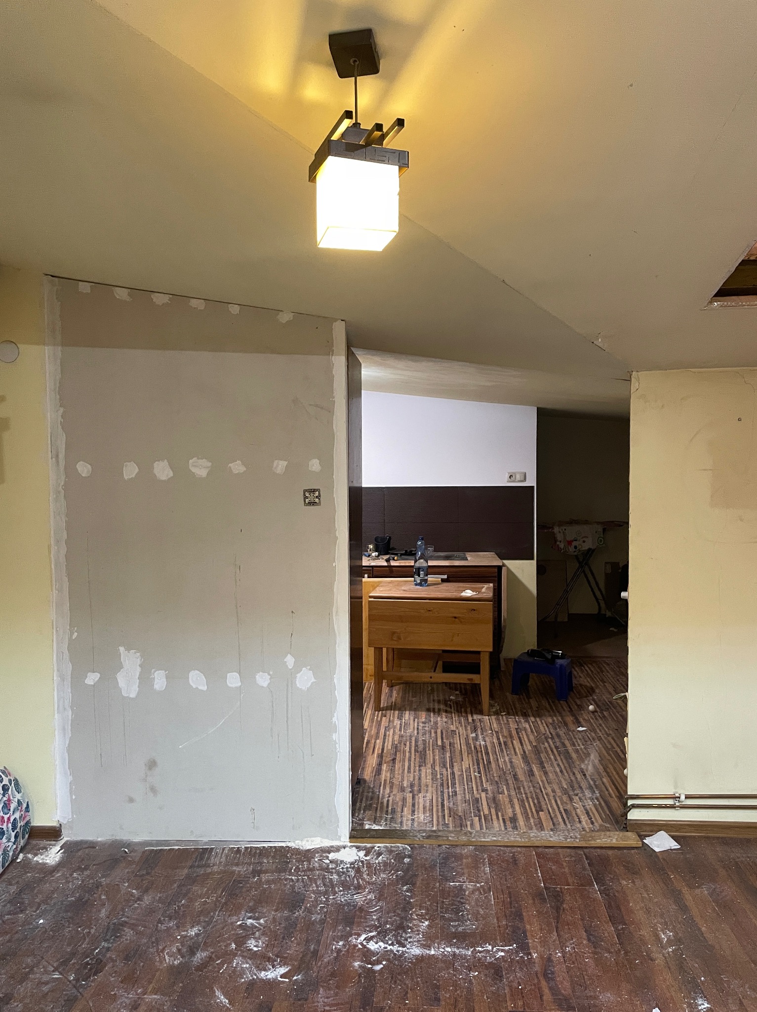 Ściana z płyty gipsowo-kartonowej po szpachlowaniu, widoczne ślady po łatach, podłoga z parkietu pokryta pyłem, w tle widoczny fragment kuchni.