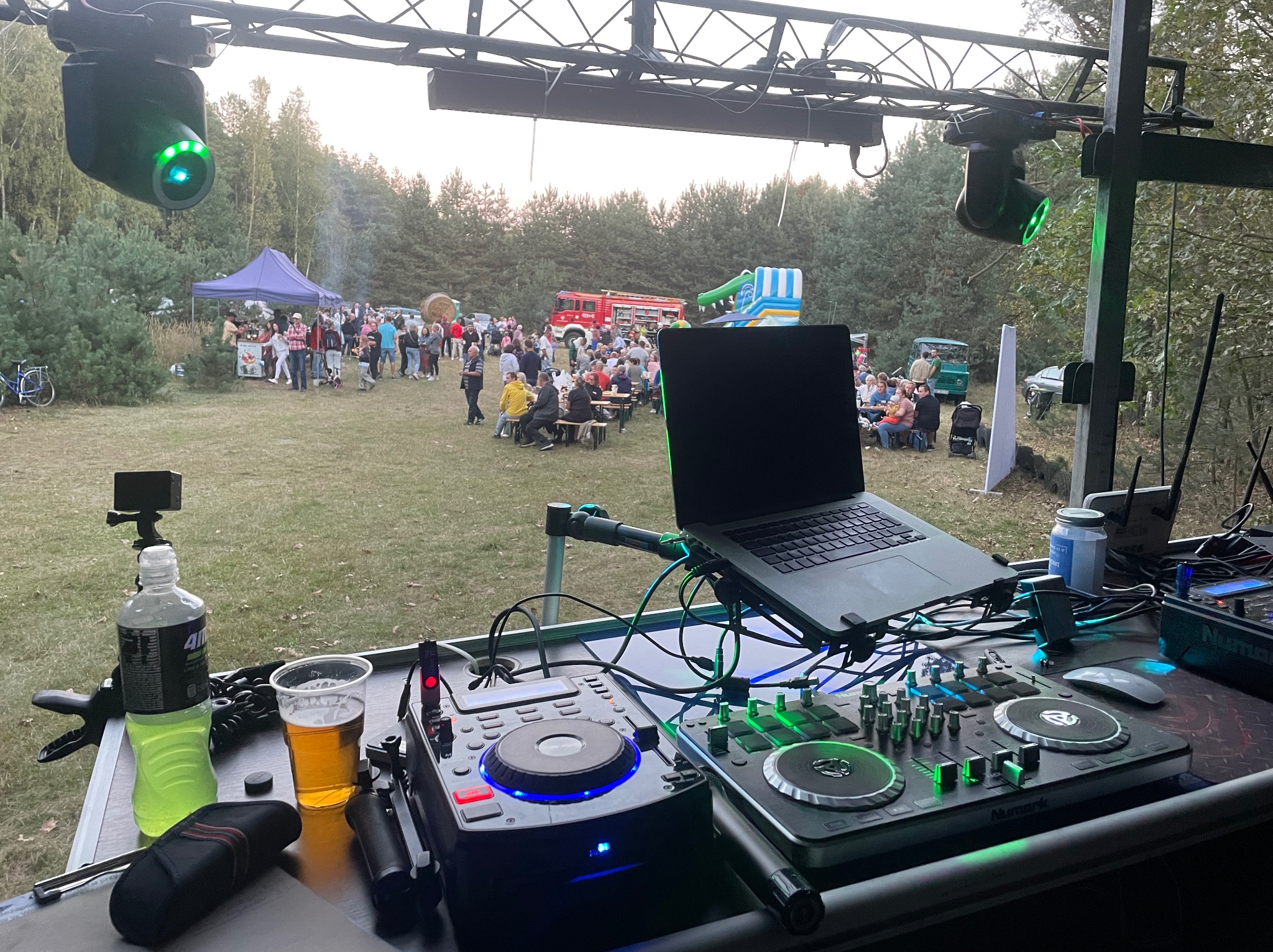 Stanowisko DJ-a na imprezie plenerowej z widocznymi gramofonami, mikserem, laptopem, napojami i oświetleniem scenicznym, w tle bawiący się ludzie, wóz strażacki i dmuchana zjeżdżalnia.