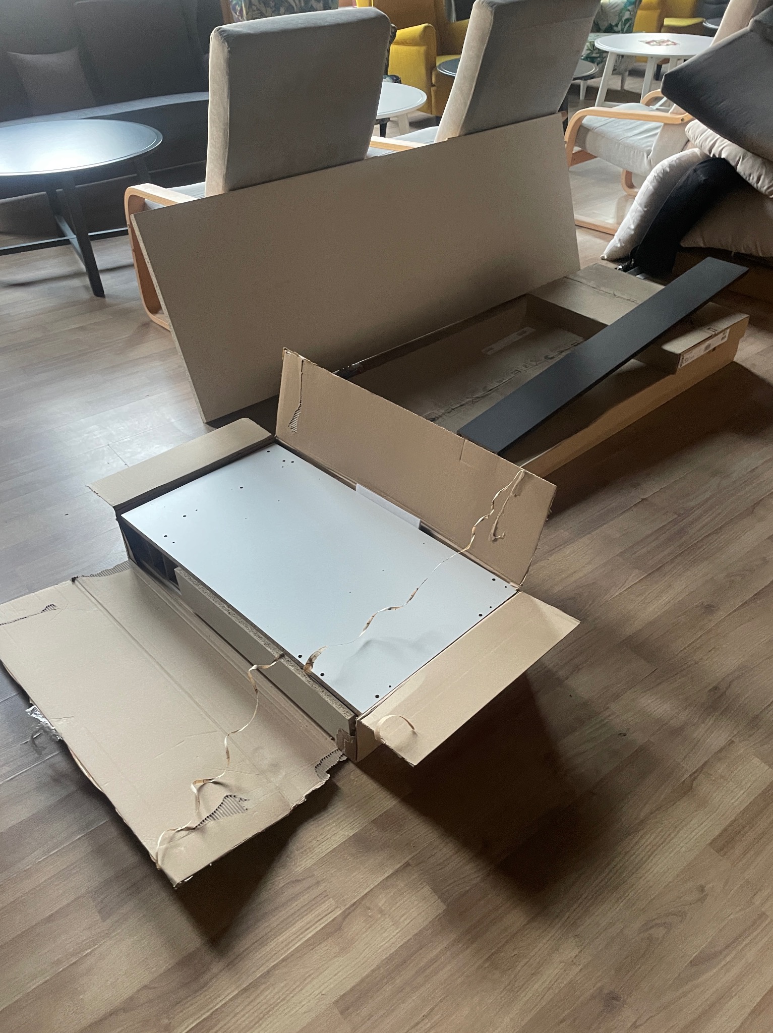 Rozpakowane elementy meblowe w kartonowych pudłach na podłodze, widoczne blaty i metalowe profile, przygotowane do montażu w pomieszczeniu z meblami w tle.